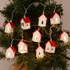Girlande aus hölzernen Weihnachtshaus-LED-Lichterketten, hängend an einer grünen Tanne.