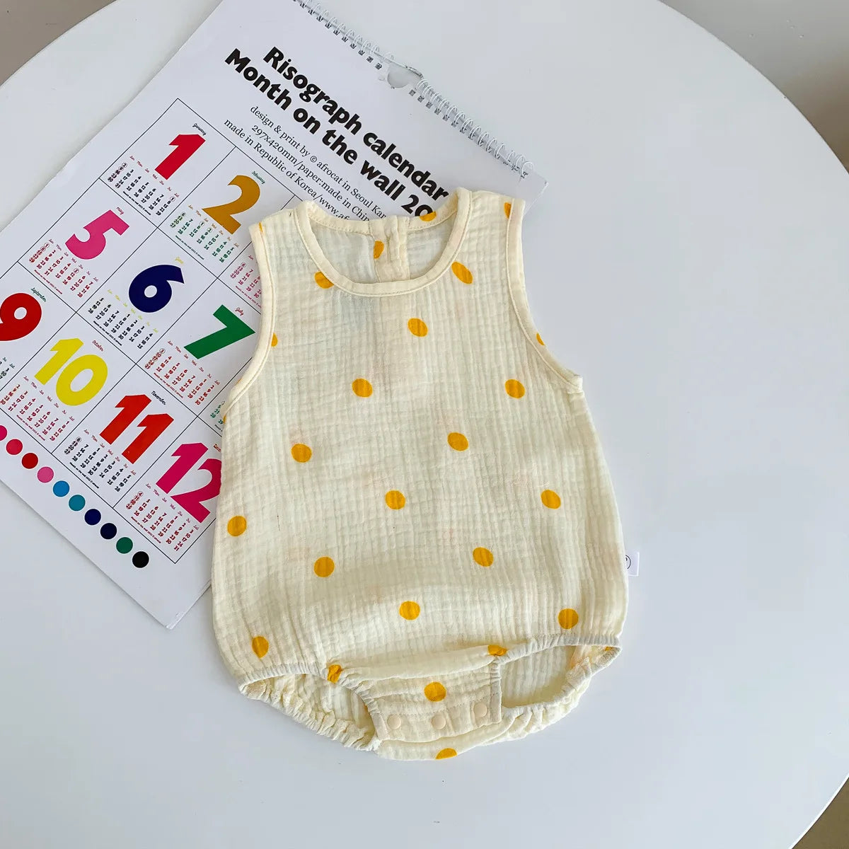 A cream-colored baby cotton sleeveless jumpsuit bodysuit with scattered orange dots, placed next to a calendar.