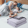 A baby is resting on a blanket next to a woman, with several folded light purple and gray throw blankets nearby.