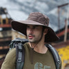 A man wearing a brown wide brim mesh bucket hat and olive green shirt with a backpack in an outdoor setting.