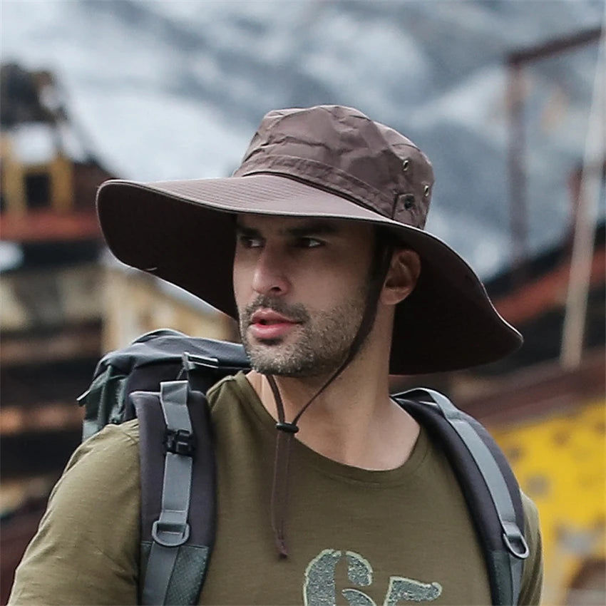 A man wearing a brown wide brim mesh bucket hat and olive green shirt with a backpack in an outdoor setting.
