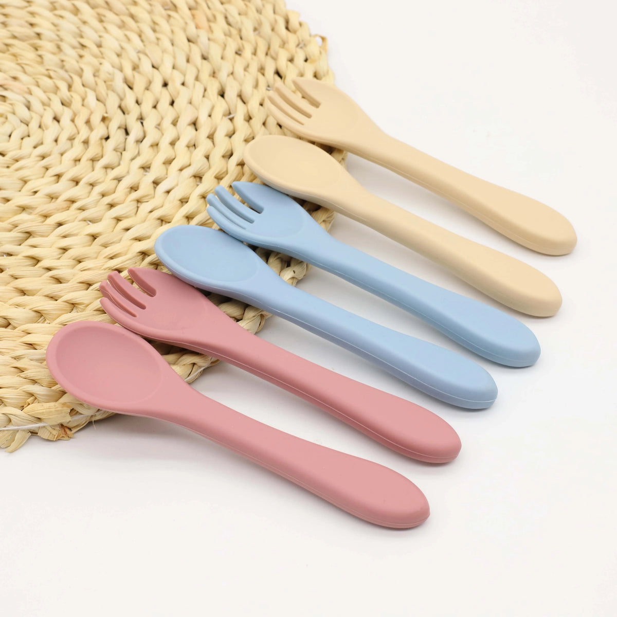 A set of five silicone baby tableware spoons and forks in pastel colors arranged on a woven mat.